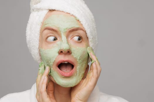 A woman with a towel on her head smiles as she applies a green face mask. She has a happy expression on her face, showing off her skincare routine