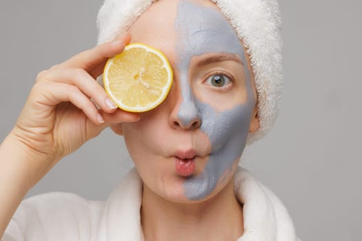 A woman with a mask on her face holding a lemon wedge in front of her mouth. She is wearing a white robe and a towel on her head. Beauty and Skin care concept