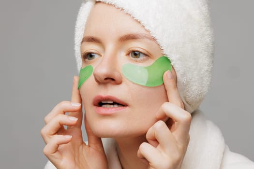 A woman with a towel on her head and a robe smiles as she applies green gel patches under her eyes facial cosmetology concept