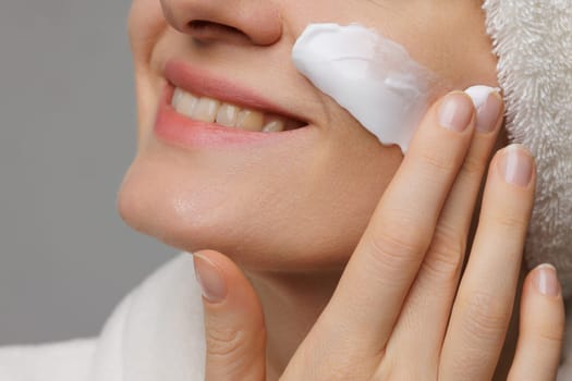 A woman applying cream to her face, smiling gently. She has a towel wrapped around her head and is in a relaxed setting, suggesting a skincare routine or spa treatment.