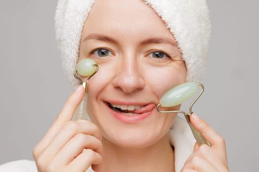 A woman with a towel on her head and a robe playfully uses jade facial rollers on her cheeks. She has a surprised expression, concept skin care facial.