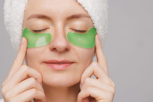 A woman with a towel on her head and a robe smiles as she applies green gel patches under her eyes facial cosmetology concept