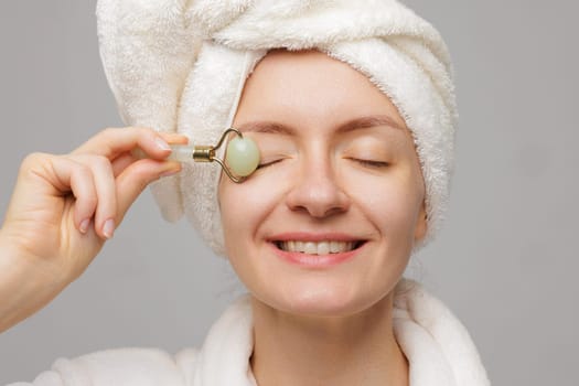 Woman with towel wrapped around head using jade roller on face smiling. Roller massaging eyes demonstrating skin care routine.