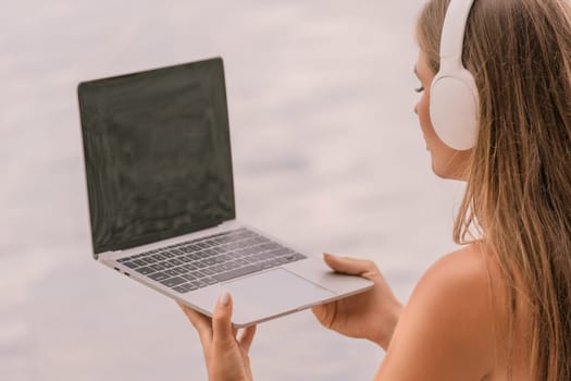 A woman is sitting on a beach with a laptop in her lap. She is wearing headphones and she is listening to music