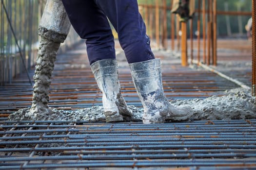Construction site worker wearing rubber boots handles a concrete pump hose while overseeing the process of pouring concrete into a reinforcement cage.