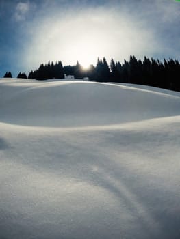 Scenic winter vista featuring mountain snowy slopes, distant trees silhouetted against the sky, sun gently breaking through the clouds, crafting a stunning portrayal of nature's beauty.