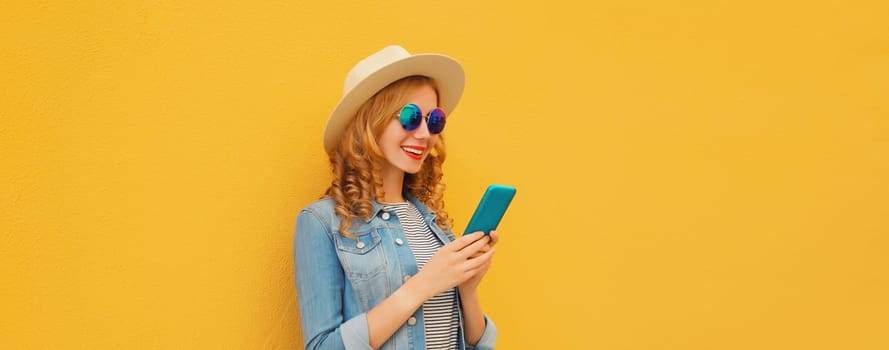 Summer portrait of happy traveler young woman with mobile phone looking at device in straw tourist hat on yellow studio background