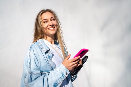 Smiling woman with blond hair holding smartphone looking at camera. Portrait of beautiful young tourist using cellphone against wall on sunny day.