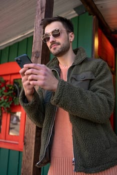 Male adult wearing sunglasses and green jacket using smartphone for sending message. Man in fashionable eyewear texting on cellphone. Modern lifestyle, connection concept.