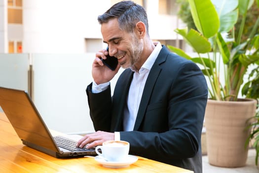 A middle-aged adult man in a suit has a business call while looking at his laptop.