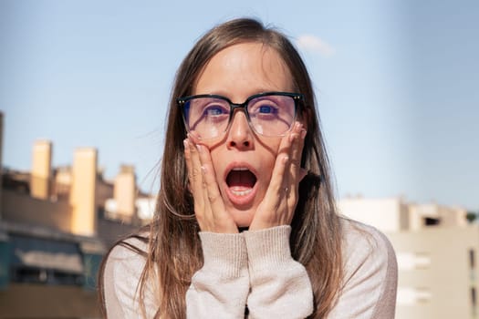 Young woman wearing glasses standing looking surprised and with her arms on her face looking at the camera.