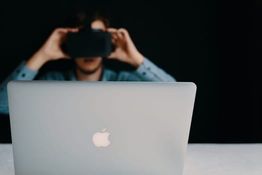 A person in a dimly lit office is using a VR headset with a laptop, creating an immersive and modern workspace. Bishkek, Kyrgyzstan - March 14, 2020