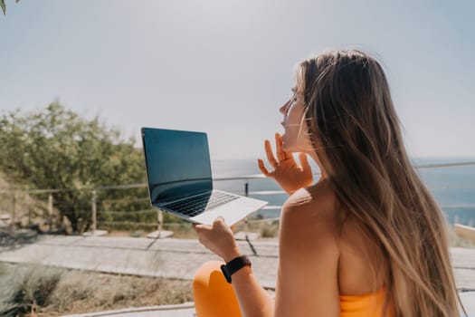A woman with long blonde hair sits outdoors and uses a laptop. She is looking at the screen and has a thoughtful expression.