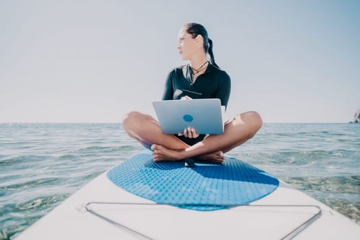Digital nomad, woman in the hat, a business woman with a laptop sits on the rocks by the sea during sunset, makes a business transaction online from a distance. Freelance, remote work on vacation.