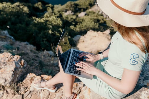 A young woman wearing a straw hat and a green dress sits on a rocky cliff, working on a laptop.