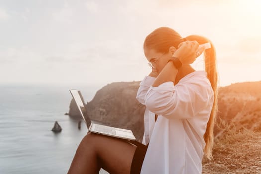 Digital nomad, woman in the hat, a business woman with a laptop sits on the rocks by the sea during sunset, makes a business transaction online from a distance. Freelance, remote work on vacation.
