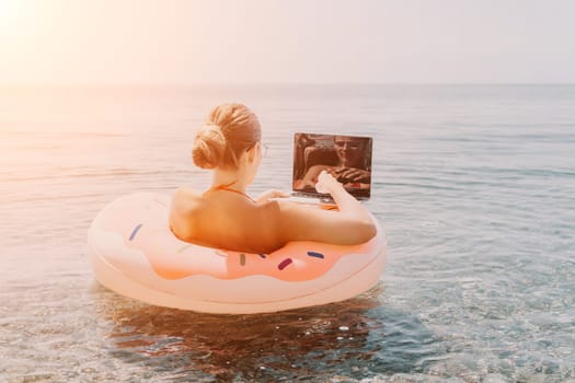 Woman freelancer works on laptop swimming in sea on pink inflatable ring. Pretty lady typing on computer while floating in the sea on inflatable donut at sunset. Freelance, remote work on vacation