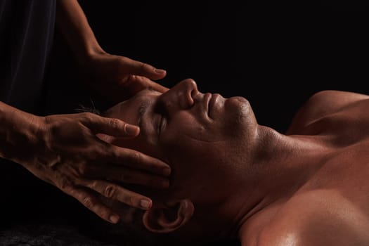 Close-up of a man receiving a facial massage against a dark background, The hands of the massage therapist are gently pressing on his forehead and temples.