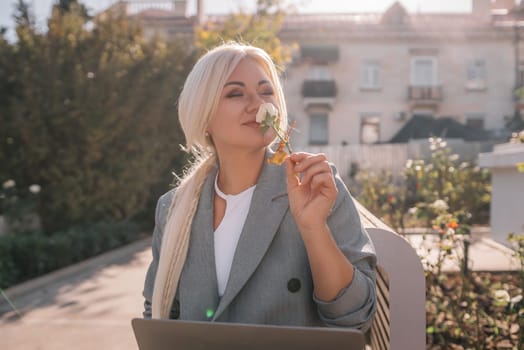 A blonde woman is sitting on a chair and eating a piece of broccoli. She is also using a laptop