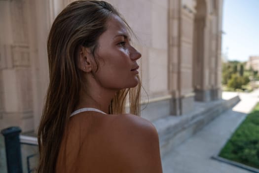A woman with long brown hair stands in front of a building, looking to the right. Her back is to the camera. She is wearing a white tank top. She is standing on a set of stairs. There is a grassy area in the background.