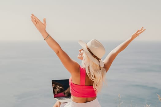 A woman is sitting on a beach with her arms outstretched, wearing a pink top and a straw hat. She is looking out at the ocean, and there is a laptop on the ground in front of her