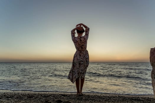 A woman in a dress stands on a beach, facing the ocean, with her arms raised above her head as the sun sets.