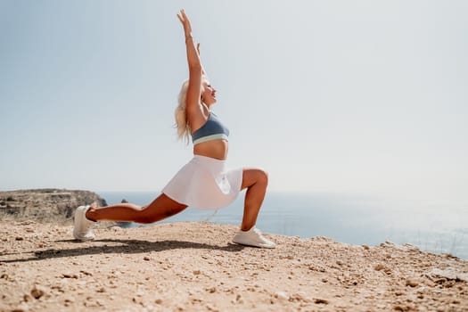 A blonde woman in a white skirt and blue sports bra performs a stretching exercise on a cliff overlooking the sea.