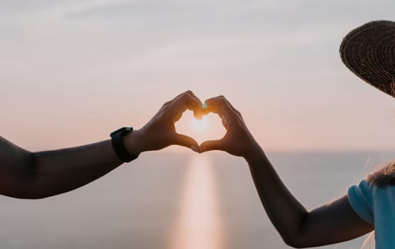 Two individuals create a heart shape with their hands as the sun sets in the background. The soft glow of the sky reflects in their silhouettes.