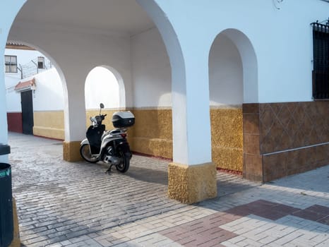 Black scooter parked under tiled archway in a European setting, with cobblestone floor. scooter parked under an archway. The scooter is black and has a storage compartment on its rear