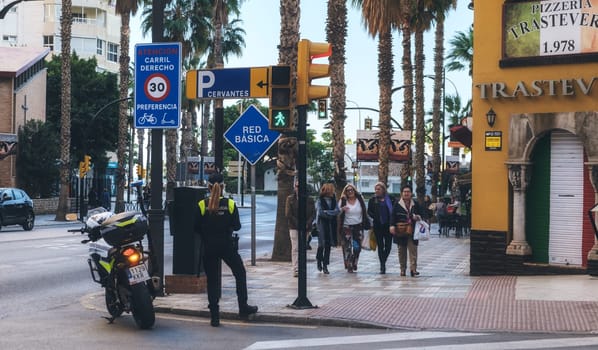 There is a traffic officer wearing a reflective vest standing near a motorcycle. directing or monitoring traffic. a group of pedestrians waiting to cross the street, and several street signs