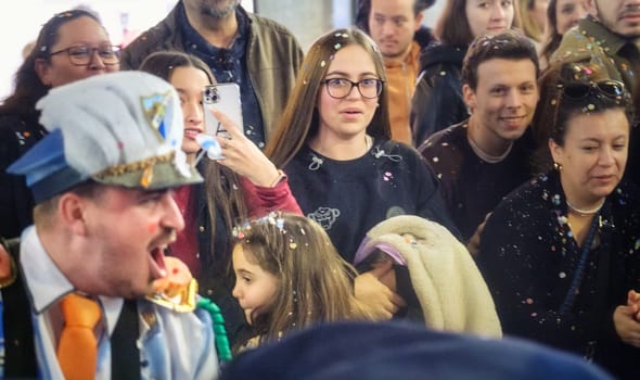 In this image, you see a group of people gathered together in what appears to be a public event or celebration. In the foreground, a person is dressed in costume, which includes a hat that looks like it might be part of a uniform, and they are making a dramatic expression with their mouth open wide...To the right of the image, there's a young woman with glasses looking toward the camera. She's wearing a black top with some pattern or design on it, and there are specks of what could be confetti in her hair and on her clothes, suggesting the festive nature of the event...Other individuals in the scene are also adorned with confetti, and some are holding items that might be part of the event or celebration. The mood overall is joyful and lively, and it seems like they are all watching or participating in some kind of performance or activity.