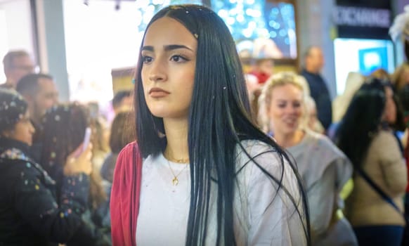 The image shows a close-up of a young woman with long dark hair and a serious expression on her face. She is wearing a white top and a gold chain necklace. In the background, there's a blurred crowd of people, suggesting that this photo was taken in a bustling, public space, possibly during an evening as the lighting seems artificial, like in a city street or a commercial area. The woman appears to be deep in thought or focused on something out of frame.