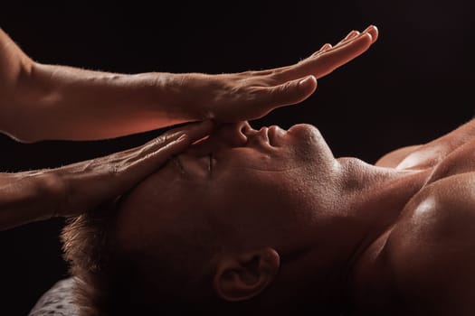 Close-up of a man receiving a facial massage against a dark background, The hands of the massage therapist are gently pressing on his forehead and temples.