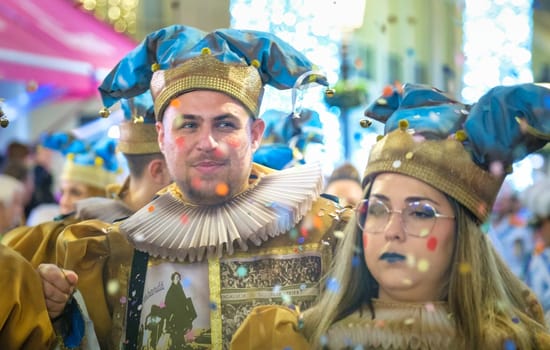 The image shows two individuals dressed in vibrant, elaborate costumes with a design reminiscent of historical royal or court attire. Their costumes feature brilliant blue and gold hues, and they are both wearing matching hats with a jester-like appearance. The person on the left sports a ruffled collar that is typical of fashion from the Elizabethan era. Both individuals have their faces decorated with colorful dots, contributing to a festive and playful look, which suggests that they may be participants in a parade, festival, or some form of costume celebration. The blurred background indicates that the photo was taken amidst bustling activity, perhaps in the middle of a lively event.