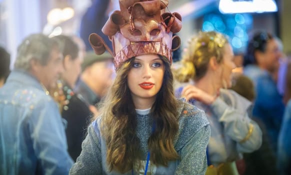 In the image, there is a person wearing an unusual hat that appears to be crafted to resemble a cake or dessert with rolling pins as decorations. The person has long hair, red lipstick, and is looking directly at the camera with a neutral expression. They are wearing a sweater or top with a shimmering texture, and behind them, there are blurred figures of other people, suggesting that the photo was taken at an event or a gathering with a fair amount of people. The lighting suggests an indoor setting with artificial illumination.
