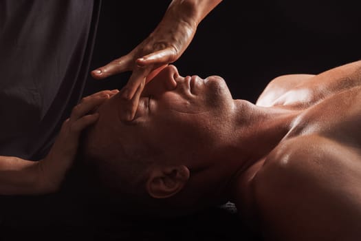 Close-up of a man receiving a facial massage against a dark background, The hands of the massage therapist are gently pressing on his forehead and temples.