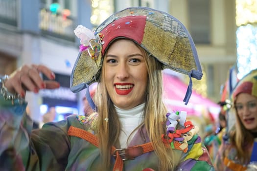 The image features a woman wearing a colorful and festive outfit, which includes a hat that appears to be elaborately designed with various patterns and bows. The material looks shiny and vivid, suggesting that it may be a costume typically worn for a celebration or a parade. She has bright red lipstick and a joyful expression on her face, suggesting she's partaking in a fun event. Her outfit includes a jacket or cloak with a patchwork of different fabrics and a white turtleneck underneath. There's a glimpse of another person behind her wearing a similarly whimsical hat, hinting that they might be a part of a group or at a themed event. The blurred background with architectural elements suggests this event is happening on a city street, possibly during a festival or carnival procession.