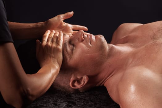 A man receiving a facial massage, lying on a dark surface. A pair of hands gently apply pressure to his forehead, promoting relaxation and wellness. The scene conveys tranquility and self-care.