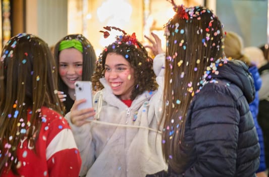 The image depicts a group of young people celebrating. At the center, there's a person with curly hair smiling broadly and looking joyous as they are surrounded by festive confetti. It appears to be a moment of celebration or a special event. To the left, another person is taking a selfie with a phone, capturing the happy moment. The people in the photo seem to be dressed for cool weather, as some are wearing jackets. The bright, warm lights and the blurriness in the background suggest that the photo was taken during the evening or at night in a lively, possibly urban environment.
