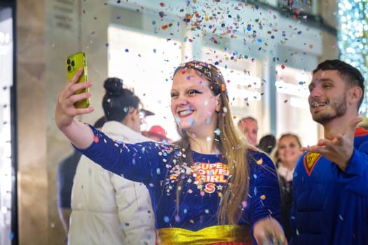 The image shows two people, a woman and a man, capturing a joyful moment with a selfie. The woman is in the foreground, taking a selfie with her smartphone, and she seems to be wearing a casual superhero-themed outfit with the text SUPER