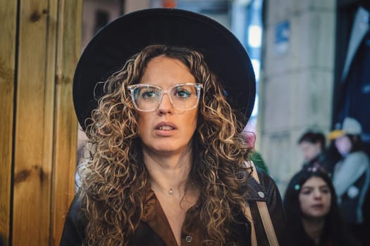 woman with curly hair wearing large, clear-framed glasses and a black hat. She has a thoughtful or focused expression on her face. She is wearing what looks like a brown coat or jacket, and behind her download photo.