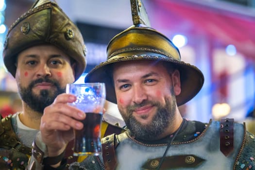 In the image, there are two individuals who appear to be dressed in costume, reminiscent of historical or fantasy armor. Both are wearing metal helmets with a peak or spike at the top, and the attire includes what looks like metallic and studded shoulder guards, suggesting a knight or warrior type of theme...The person on the right is smiling and holding up a glass with a dark liquid, possibly beer, in a toasting gesture. The other individual is also smiling but isn't directly engaging with the camera. They both have red marks on their cheeks, which could be part of the costume effect or some kind of celebration markings. The setting appears to be a festive or social environment, likely a place where dressing up and enjoying beverages is part of the event's atmosphere.