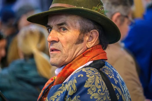 older man from the side view wearing a distinctive outfit. He has on a hat, He wears a patterned scarf around his neck, and his jacket is ornate. The man has a stern expression on his face