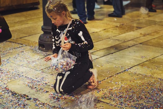 The image shows a young girl kneeling on the floor surrounded by colorful confetti. She appears to be holding onto a clear plastic bag that contains more confetti, suggesting that she may have been collecting it or that the bag has spilled. Her focus and the way she's interacting with the confetti give the impression that she is either playing or cleaning up. The environment looks like an indoor venue, possibly after a celebration or an event where confetti was used. The scattered confetti adds a vibrant touch to the image, reflecting a festive or celebratory atmosphere.