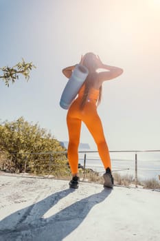 A woman wearing an orange yoga outfit with a blue yoga mat on her back is standing on a concrete surface, looking out at the sea.