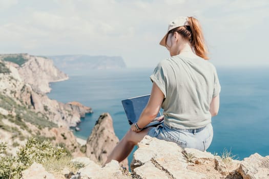 Digital nomad, woman in the hat, a business woman with a laptop sits on the rocks by the sea during sunset, makes a business transaction online from a distance. Freelance, remote work on vacation.