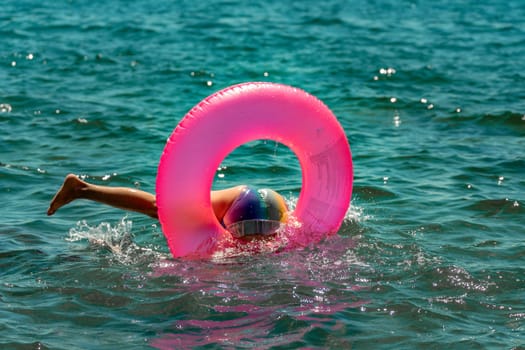 A child wearing a blue and white hat floats on a pink inflatable ring in the blue ocean. They are upside down and submerged.