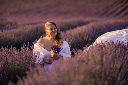 Woman poses in lavender field at sunset. Happy woman in white dress holds lavender bouquet. Aromatherapy concept, lavender oil, photo session in lavender.