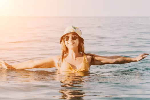 A woman in a yellow bikini and bucket hat floats in the ocean with her arms outstretched.