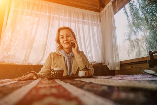 A woman sits at a table in a wooden gazebo, talking on a cell phone. Sunlight streams through the window behind her.